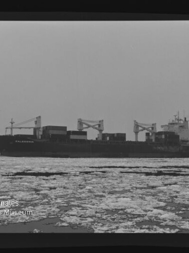 Frachtschiff mit Ladekränen fährt auf ruhiger, teilweise zugefrorener See, Vordergrund mit Treibeis und Museumslogo