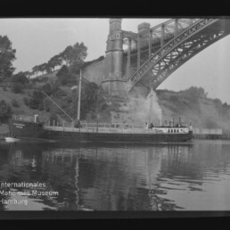 Frachtschiff auf ruhigem Fluss mit Spiegelung, dampfender Schornstein unter einer großen Stahlbogenbrücke, bewaldetes Ufer.