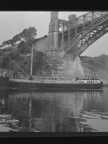 Frachtschiff auf ruhigem Fluss mit Spiegelung, dampfender Schornstein unter einer großen Stahlbogenbrücke, bewaldetes Ufer.
