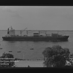 Frachtschiff mit Ladekränen auf dem Wasser, Uferbäume und kleine Boote; Logo Internationales Maritimes Museum Hamburg.