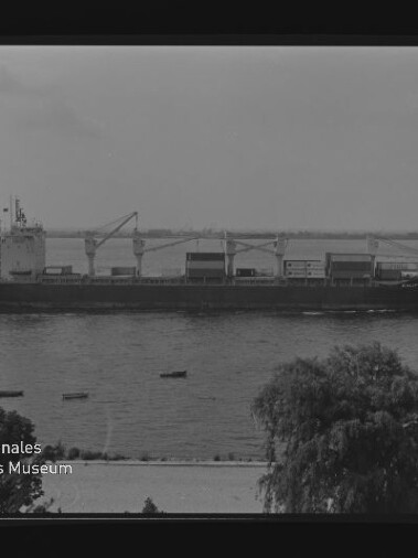 Frachtschiff mit Ladekränen auf dem Wasser, Uferbäume und kleine Boote; Logo Internationales Maritimes Museum Hamburg.