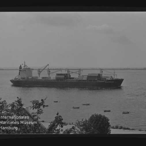 Frachtschiff mit Ladekränen und Containern auf ruhiger See, Blick vom Ufer mit Bäumen im Vordergrund und Museumslogo unten links