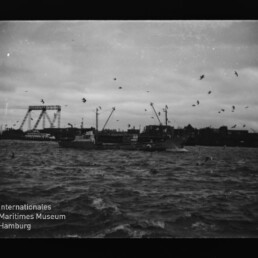 Schwarzweiß-Hafen mit Frachtschiffen, zahlreichen Möwen über aufgewühlter See und einer markanten Klappbrücke im Hintergrund.