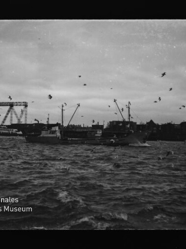 Schwarzweiß-Hafen mit Frachtschiffen, zahlreichen Möwen über aufgewühlter See und einer markanten Klappbrücke im Hintergrund.