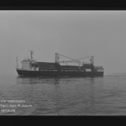 Graustufenfoto eines Frachtschiffs mit zwei Kränen auf ruhiger See; Museumslogo unten links.