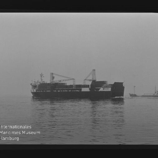 Frachtschiff mit zwei Ladekränen auf ruhigem Meer, rechts ein kleiner Schlepper und Wasserzeichen des Maritimen Museums.