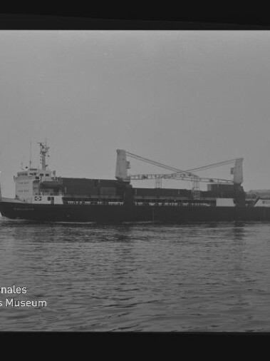 Frachtschiff mit zwei Deckkränen auf ruhiger See, kleines Begleitschiff links und Museumslogo unten links.