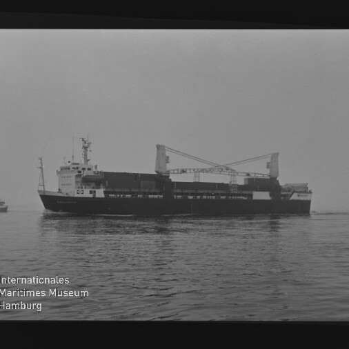 Frachtschiff mit zwei Deckkränen auf ruhiger See, kleines Begleitschiff links und Museumslogo unten links.