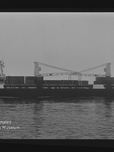 Schwarzweiß-Foto eines Frachtschiffs mit zwei Kranauslegern und Containern auf ruhiger See, Museumslogo unten links.