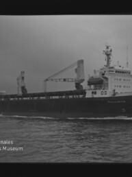 Schwarzweißfoto eines Frachtschiffs mit zwei Deckkränen und vorn liegender Brücke, auf ruhiger See vor bewölktem Himmel; Museumslogo unten links.