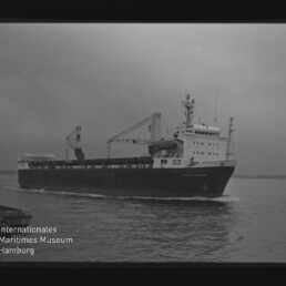 Schwarzweißaufnahme eines Frachtschiffs mit Ladekranen, das auf ruhiger See vorbeifährt; Museumssignet unten links.