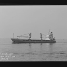Frachtschiff mit zwei Ladekränen auf ruhiger See, seitliche Schwarzweißaufnahme mit flachem Horizont.