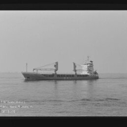 Frachtschiff mit zwei Ladekränen auf ruhiger See, Schwarzweißfoto mit Filmrand und Schriftzug AGFA APX 100.
