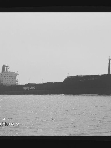Seitliche Aufnahme eines Frachtschiffs mit Hapag-Lloyd-Logo auf ruhiger See in schwarzweiß; Museumslogo unten links.