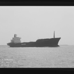 Schwarz-weiß-Foto eines Frachtschiffs in Seitenansicht, das ruhig auf dem Meer vor einem flachen Horizont fährt.