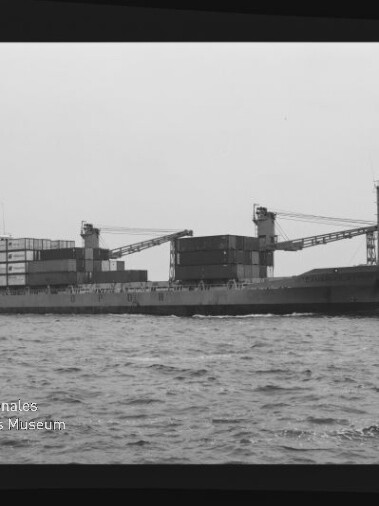 Containerschiff mit aufgestapelten Containern und zwei Ladekränen fährt auf ruhiger See; schwarzweiß mit Museumslogo.