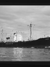 Frachtschiff Candia mit Ladekränen und mittigem Aufbau fährt auf ruhigem Meer; Museum-Logo unten links.