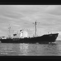 Frachtschiff Candia mit Ladekränen und mittigem Aufbau fährt auf ruhigem Meer; Museum-Logo unten links.