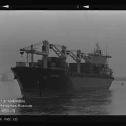 Containerschiff CAMBIA mit Ladekränen und gestapelten Containern fährt auf ruhigem Wasser; Schwarzweiß-Filmrand sichtbar.
