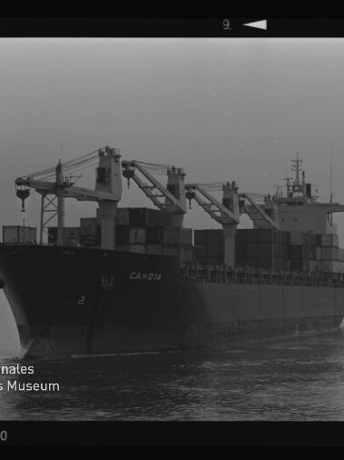 Containerschiff CAMBIA mit Ladekränen und gestapelten Containern fährt auf ruhigem Wasser; Schwarzweiß-Filmrand sichtbar.