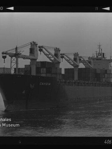 Schwarzweißfoto eines Containerschiffs mit der Aufschrift CAMBIA, sichtbaren Bordkränen und gestapelten Containern, Museumswasserzeichen links unten.