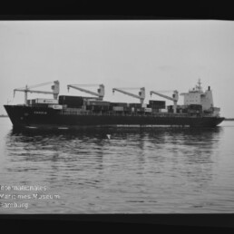 Frachtschiff mit vier Deckskranen und gestapelten Containern, ruhig auf spiegelndem Wasser gelegen.