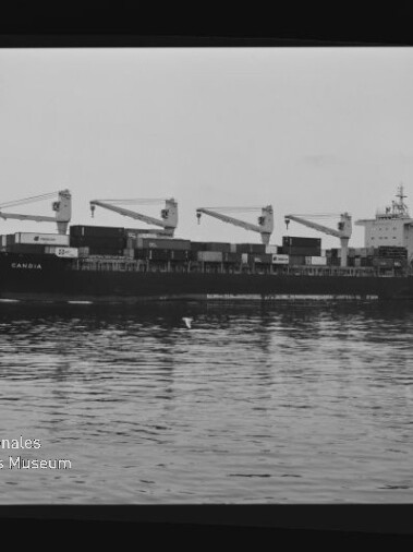 Frachtschiff mit vier Deckskranen und gestapelten Containern, ruhig auf spiegelndem Wasser gelegen.