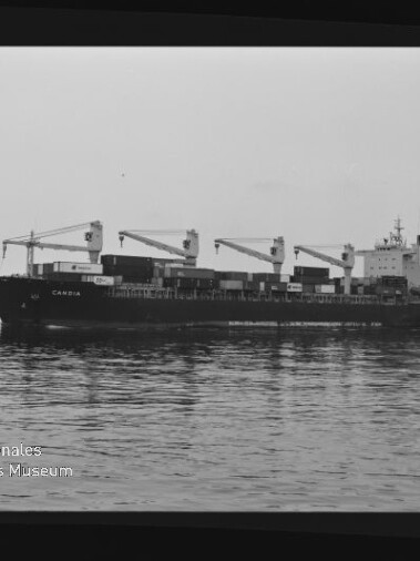 Frachtschiff mit vier Ladekränen und Containern fährt über spiegelndes Wasser; schwarz-weiß-Aufnahme.