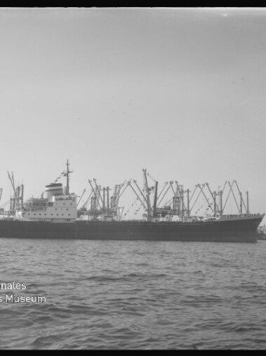 Großes Frachtschiff mit zahlreichen Ladekränen vor einem Hafen, ruhige See im Vordergrund und Museumssignet unten links.