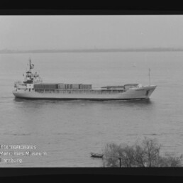 Schwarzweißfoto eines Frachtschiffs mit Containern auf ruhigem Wasser, im Vordergrund Uferbüschen und Museum-Wasserzeichen