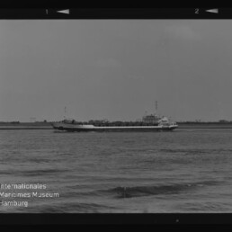 Schwarzweiß-Foto eines langen Frachtschiffs auf ruhigem Wasser vor flachem Ufer; Filmrand und Museumstext links unten sichtbar.