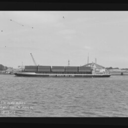Frachtschiff mit aufgestapelten Containern und Schrift GEEST NORTH SEA LINE, vor Hafen mit Kränen, schwarz-weiß Aufnahme.