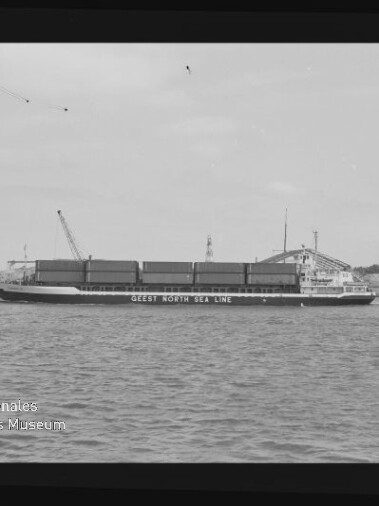 Frachtschiff mit aufgestapelten Containern und Schrift GEEST NORTH SEA LINE, vor Hafen mit Kränen, schwarz-weiß Aufnahme.