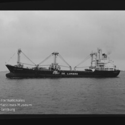 Schwarzweiß-Foto eines Frachtschiffs mit der Aufschrift PORT DE LUMIERE, Ladekränen und Heckaufbauten auf ruhiger See, Museumslogo links unten.