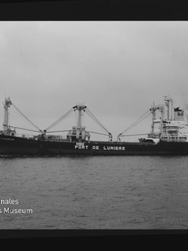 Schwarzweiß-Foto eines Frachtschiffs mit der Aufschrift PORT DE LUMIERE, Ladekränen und Heckaufbauten auf ruhiger See, Museumslogo links unten.