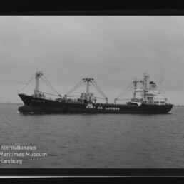 Schwarzweißaufnahme eines Frachtschiffs mit mehreren Ladekränen und der Rumpfbeschriftung PORT DE LUMIERE auf ruhiger See.