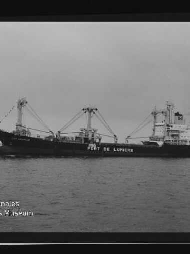 Schwarzweißaufnahme eines Frachtschiffs mit mehreren Ladekränen und der Rumpfbeschriftung PORT DE LUMIERE auf ruhiger See.