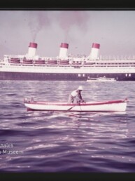 Person rudert in kleinem Ruderboot vor einem großen Passagierdampfer mit drei Schornsteinen im Hafen.