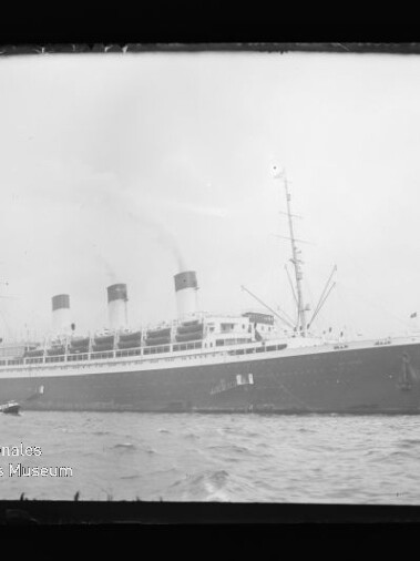Großes historisches Passagierschiff mit drei Schornsteinen im Hafen, Wasser im Vordergrund und Glockenturm im Hintergrund.