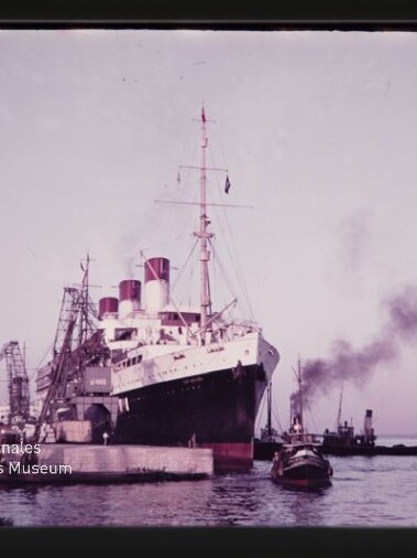 Großer Ozeandampfer mit drei roten Schornsteinen am Kai neben Kränen, Schlepper mit Rauch und Museums‑Wasserzeichen