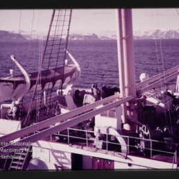 Schiffsdeck mit Rettungsbooten, Masten und Crew, Blick aufs Meer und schneebedeckte Berge; Wasserzeichen Maritimes Museum