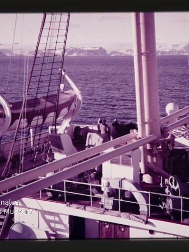 Schiffsdeck mit Rettungsbooten, Masten und Crew, Blick aufs Meer und schneebedeckte Berge; Wasserzeichen Maritimes Museum