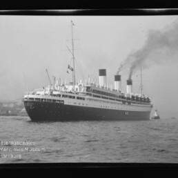 Großer vintage Ozeandampfer mit drei rauchenden Schornsteinen, begleitet von einem Schlepper vor einem Hafen.