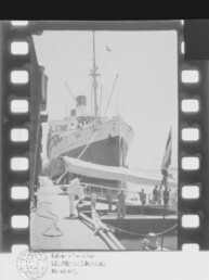 Großes Passagierschiff mit markantem Bug und Schornstein am Kai, Menschen und Beiboot am Steg.