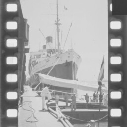 Großes Passagierschiff mit markantem Bug und Schornstein am Kai, Menschen und Beiboot am Steg.
