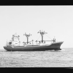Frachtschiff mit mehreren Ladekränen auf ruhiger See, fährt mit Bugwelle; Schwarzweißaufnahme mit Museumswasserzeichen.