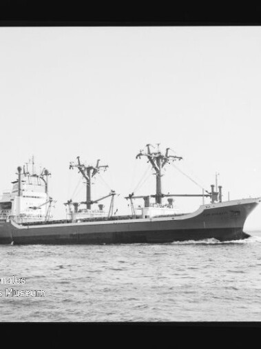 Frachtschiff mit mehreren Ladekränen auf ruhiger See, fährt mit Bugwelle; Schwarzweißaufnahme mit Museumswasserzeichen.