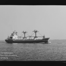 Frachtschiff mit drei Ladekränen fährt seitlich auf ruhiger See, schwarzweißes Foto mit Wasserzeichen des Maritimen Museums.