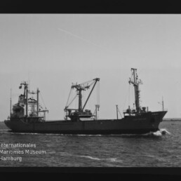Frachtschiff mit drei Ladekränen fährt auf ruhiger See, dunkler Filmrand und Museumslogo am linken unteren Bildrand sichtbar.