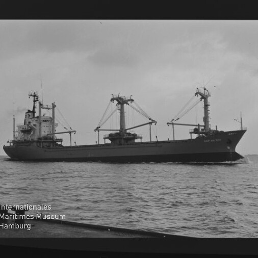 Schwarzweißfoto eines Frachtschiffs mit drei Ladekränen auf ruhiger See, unten links Logo Museum Hamburg